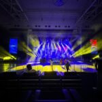 Hariharan performing live on stage with a microphone and band, in front of a large crowd and colorful LED screen.