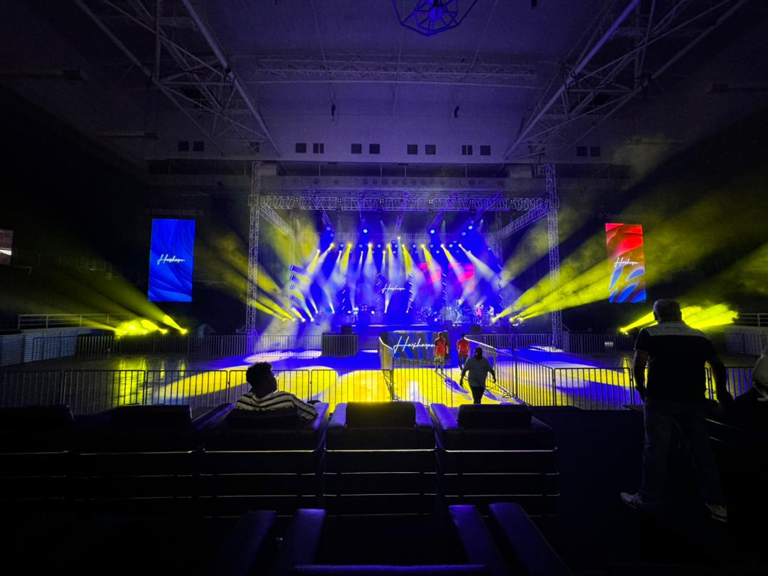 Hariharan performing live on stage with a microphone and band, in front of a large crowd and colorful LED screen.