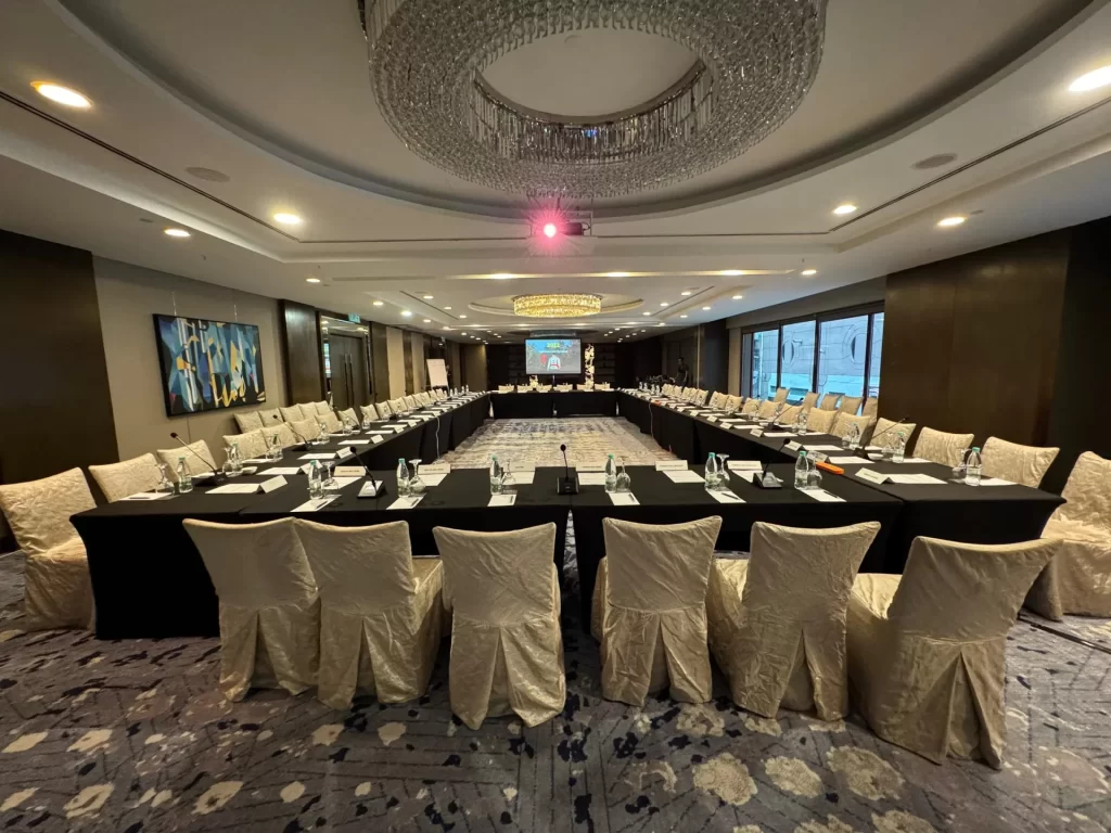 The image shows the meeting location, with gooseneck mics at every participant's desk, sound system and also projector showing slides at the end of the room.